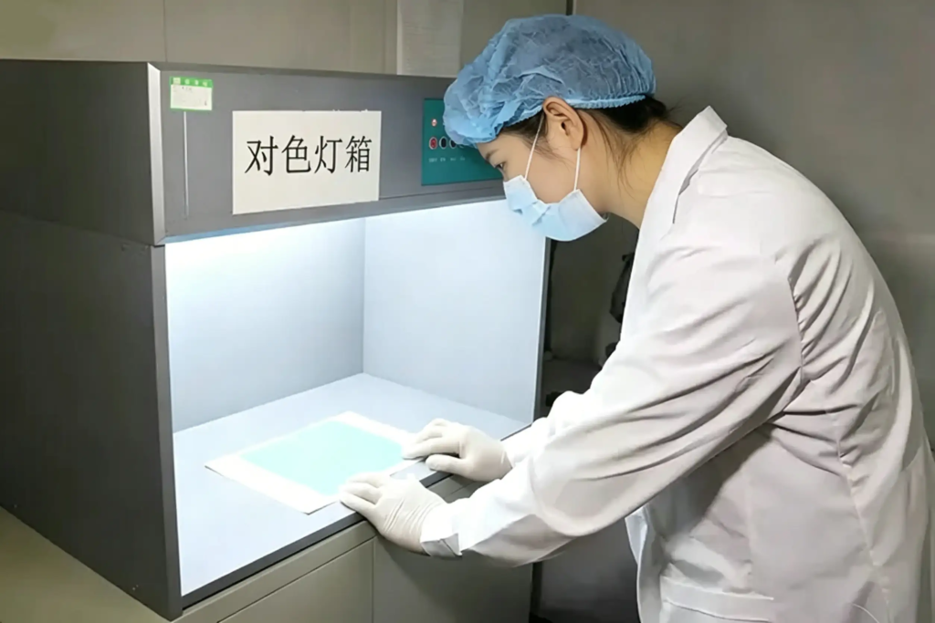 Female inspector checking color consistency of non-woven fabric against standard sample in color matching light box at Takewind non-woven factory lab