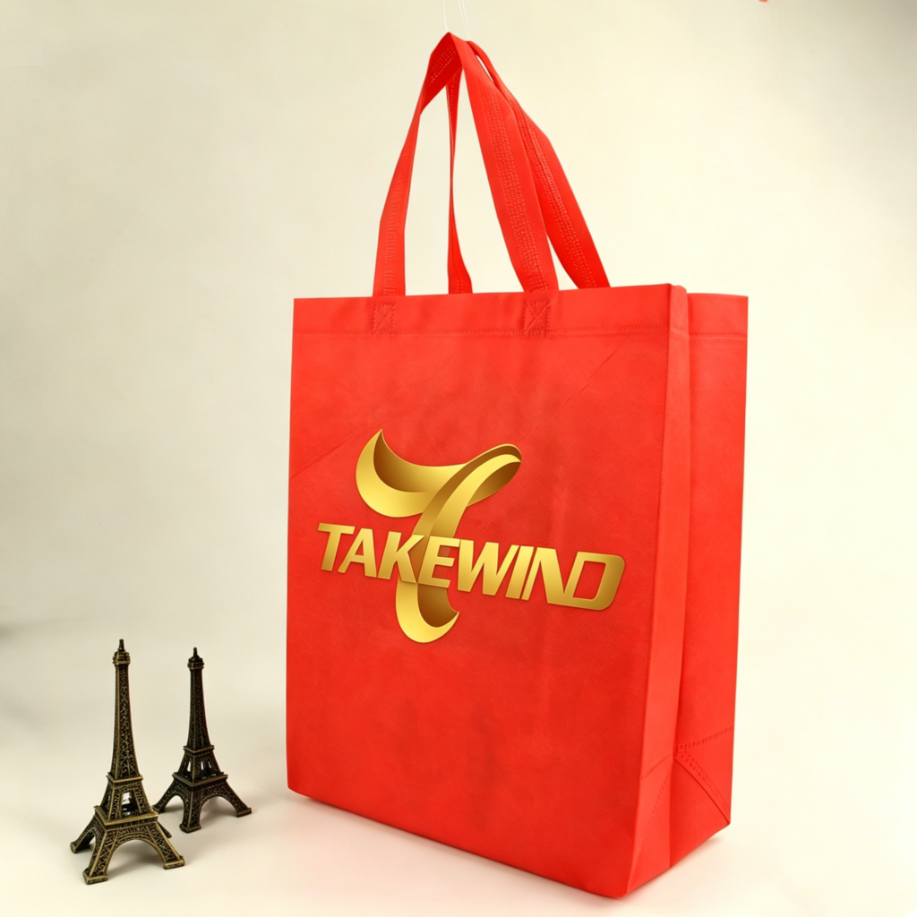 A red non-woven eco-friendly shopping bag, featuring a gold-printed TAKEWIND company logo on its front, stands upright against a clean beige background, highlighting the bag's non-woven texture, handle stitching, and brand identity.