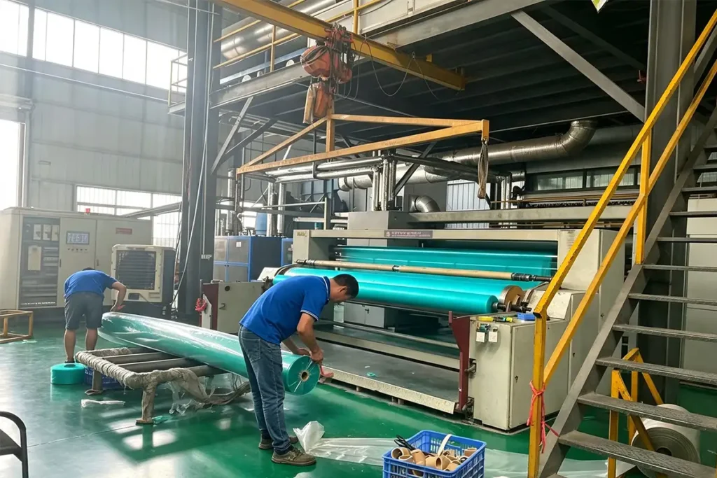 Workers finishing and packing large green geotextile rolls on industrial winding machine in clean factory workshop