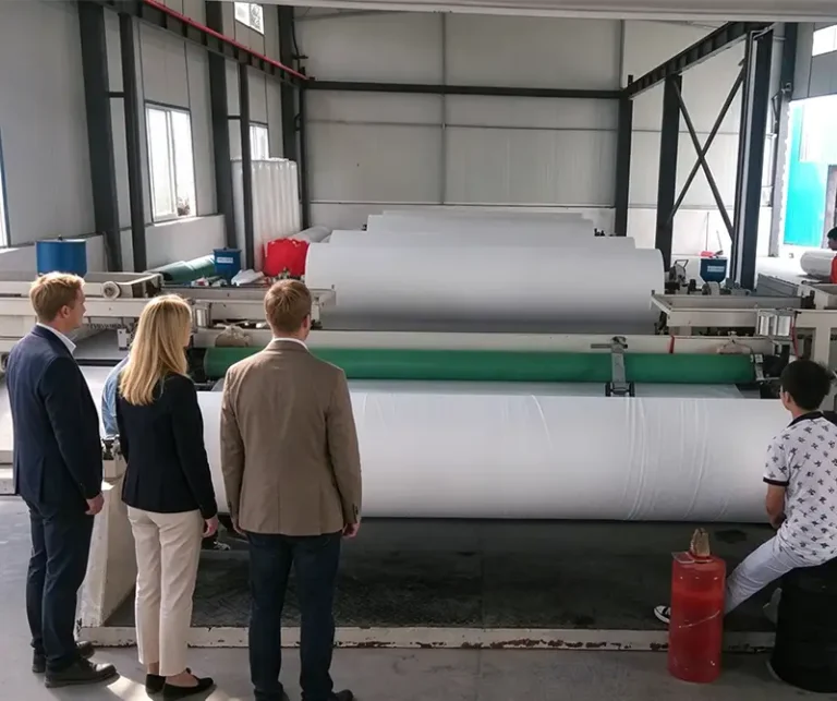 Two men and one female British customer standing back view, visiting and inspecting agricultural non-woven fabric production line inside Takewind non-woven textile manufacturing workshop, factory production equipment and large white non-woven fabric rolls in view